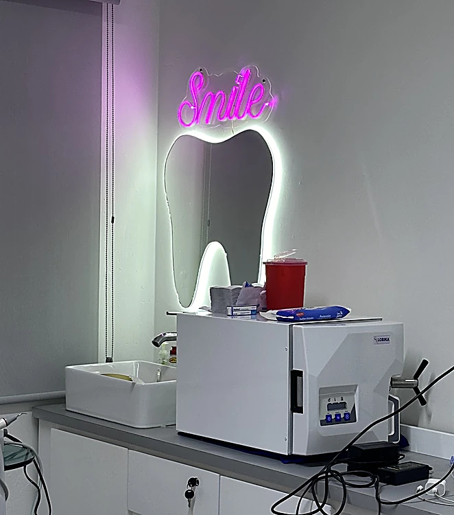 Consultorio de Dentader: espejo en forma de diente con letrero de neón rosa que dice Smile, lavabo y esterilizador